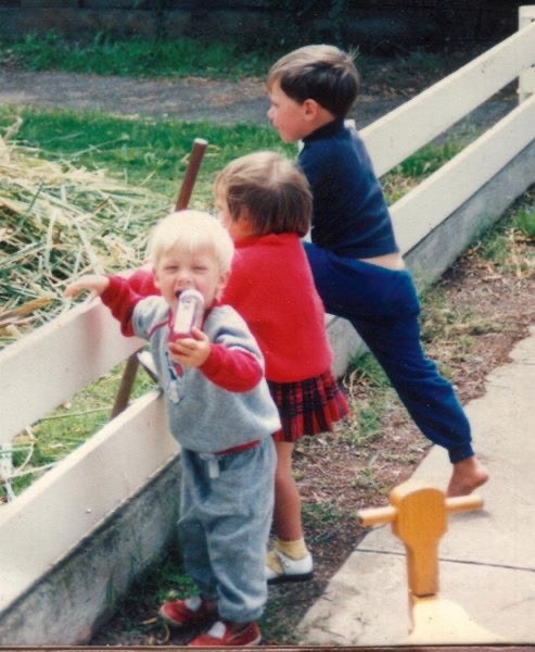 Three children beside a low garden fence with straw behind them; original date and identities unknown
