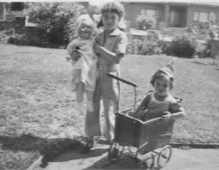 Black-and-white photograph of two children standing beside a third child in a toy pram on a lawn; original date and identities unknown